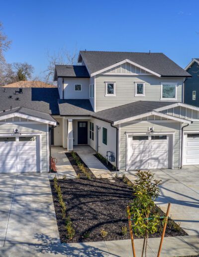 Two-story modern gray house with a large driveway and two separate garages, surrounded by a neat garden and wooden fence, under a clear blue sky.