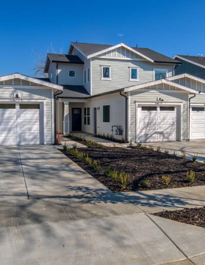 A modern duplex house with white siding, two garages, and a shared driveway. A small garden area is in front, and a wooden fence runs along the side. Clear blue sky in the background.