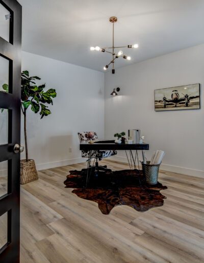 Modern home office with double glass doors, a black desk, a cowhide rug, and a plant. A minimalist chandelier hangs from the ceiling.