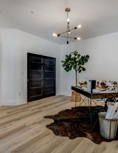 A modern home office with a black desk, chair, cowhide rug, and potted plant. Double doors on the left, a black door in the center, and a ceiling light fixture. Neutral wall and floor tones.