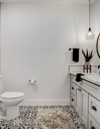 A modern bathroom with a white toilet, black and white patterned tile floor, dual sink vanity, round mirror, and pendant lights. A plant and toiletries are on the countertop.