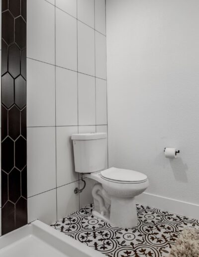 A bathroom with a white toilet, white tiled walls, decorative patterned floor tiles, a shower area, and a toilet paper holder.