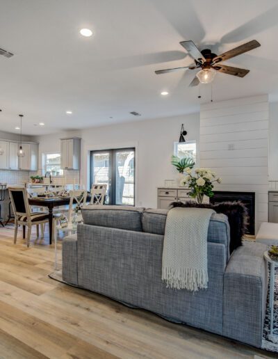 Open-concept living room and kitchen with a gray sofa, wooden dining table, and ceiling fan. Decor includes plants, wall art, and a shiplap panel accent on the fireplace.