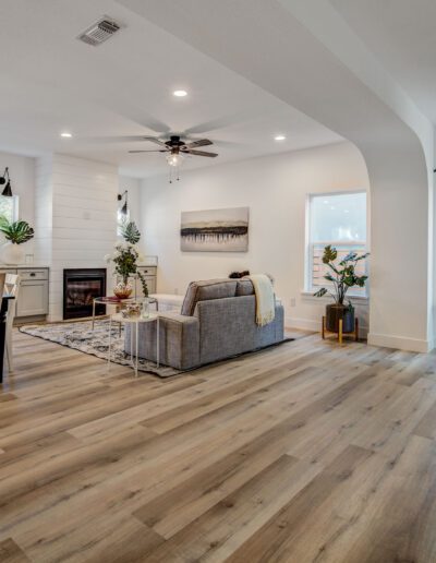 Open-concept living and dining area with wood flooring, a ceiling fan, and large windows. Features a dining table, sofa, fireplace, and minimalist decor.