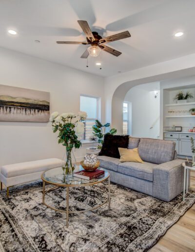 A modern living room with a gray sofa, patterned rug, ceiling fan, and wall art. There are flowers on the coffee table and shelves with decorative items in the background.