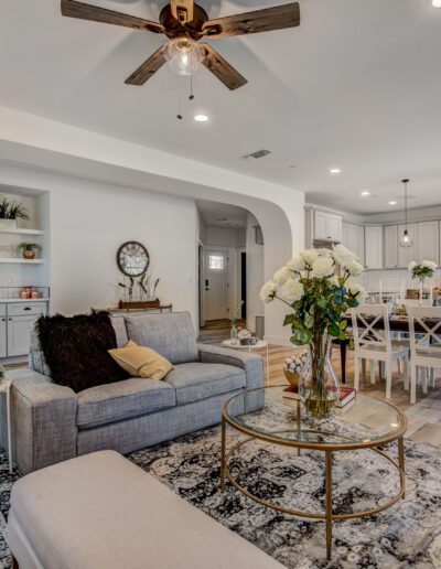 A modern open-concept living room and kitchen with a gray sofa, glass coffee table, dining table, and ceiling fan. Decor includes plants and a patterned rug.