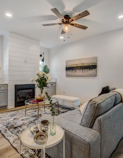 A modern living room with a gray couch, a fireplace, two windows, and a ceiling fan. Decor includes plants, a painting, and a patterned rug on light wood flooring.