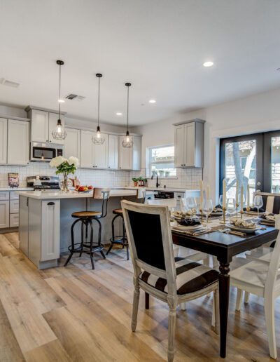 Modern kitchen and dining area with pendant lights, a central island, stools, a set dining table, and wooden flooring. Cabinets are light gray, and a sliding door opens to a patio.