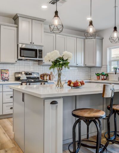 Modern kitchen with white cabinets, a large island with wooden stools, pendant lights, and hardwood floors. A vase with flowers and apples is on the island.