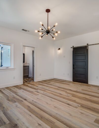 A modern, empty room with wooden flooring, a black barn door, a geometric light fixture, and two windows. Another door leads to an adjacent room.