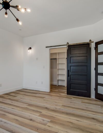 Empty room with wood flooring, black barn-style doors, and a ceiling light fixture. An open door leads to a bathroom with a vanity and mirror. A closet with shelves is visible.