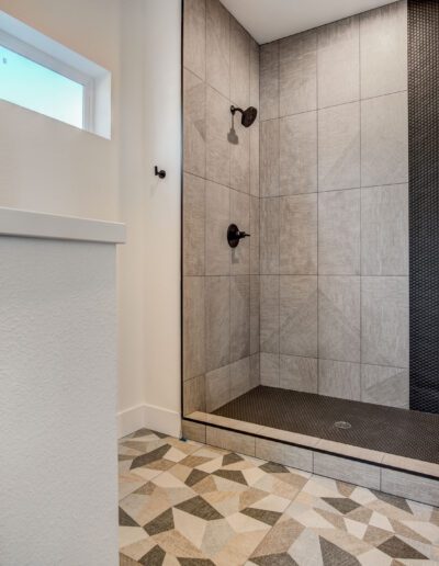 A modern bathroom with a glass-enclosed shower featuring gray tiles and black fixtures. The floor has a geometric patterned design.