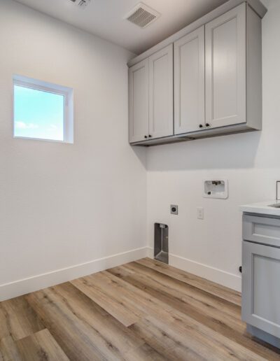 A small laundry room with light gray cabinets, a sink, a faucet, and wood flooring. A small window is on the left wall.