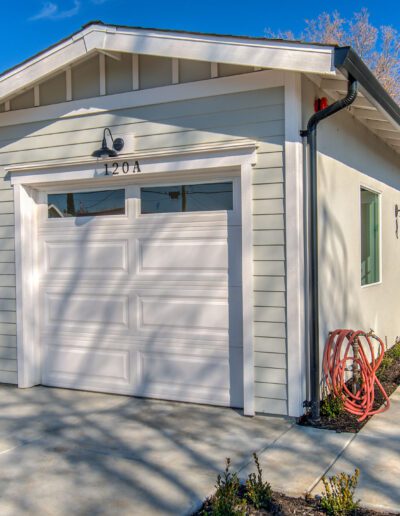 A white garage with a closed door, concrete driveway, coiled red hose, and wooden fence on the left, under a clear blue sky.