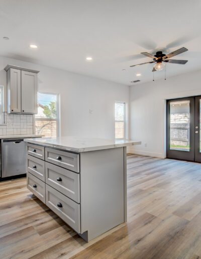 A modern kitchen with gray cabinets, a center island, and stainless steel appliances. The room features wooden floors, recessed lighting, and a ceiling fan, with glass doors leading outside.