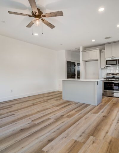 Modern open-plan kitchen and living area with wooden flooring, light grey cabinets, stainless steel appliances, and an overhead ceiling fan.