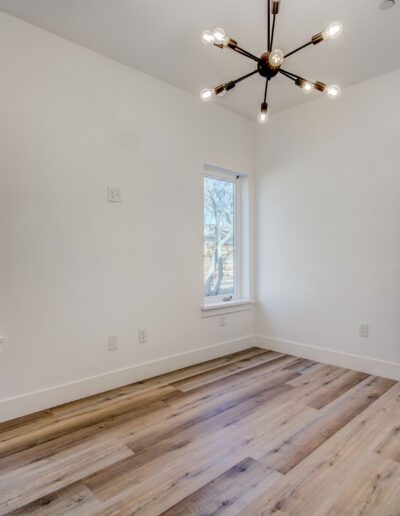 A small, empty room with light wood flooring, two windows, a modern light fixture, and an open door leading to a bathroom with black-and-white floor tiles.