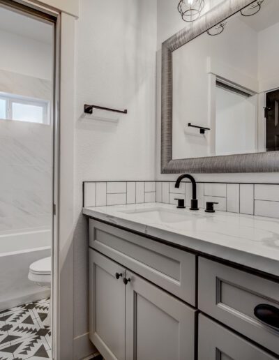 Modern bathroom with a light gray vanity, black fixtures, white tile backsplash, large mirror, and patterned black-and-white floor. A bathtub with a tile wall and small window is in the background.