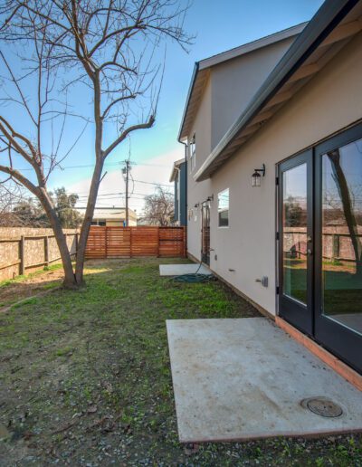 Backyard view with a tree, a wooden fence, and modern house featuring sliding glass doors. Sparse grass and concrete slabs on the ground.