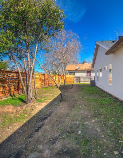 A backyard with a fence, a few trees, a grassy area, and a house on the right under a clear blue sky.