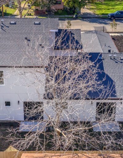 Aerial view of a two-story house with a grey roof, visible tree without leaves in the backyard, surrounded by a wooden fence, and a street in the background.