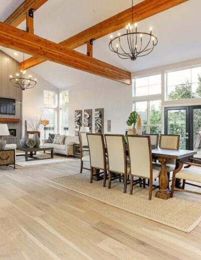 Spacious open-concept living and dining area with wooden beams, large windows, modern chandeliers, and light hardwood floors.