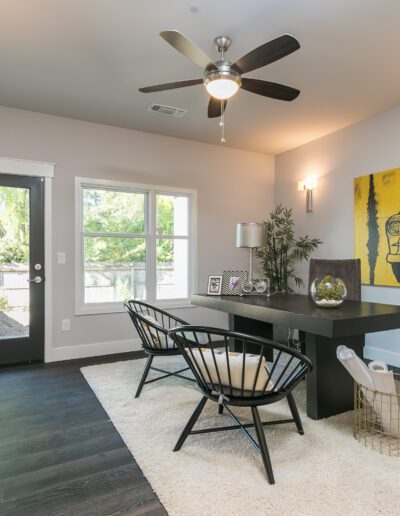 A modern home office with a black desk, two chairs, a yellow car-themed art piece, and plants, under a ceiling fan. Natural light enters through a window and door.