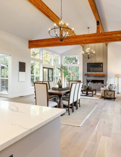 Spacious living and dining area with high ceilings, wooden beams, and large windows. Features a fireplace, modern chandeliers, and a view of a sunroom through glass doors.