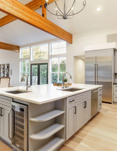 Spacious kitchen with light wood floors, gray cabinets, dual islands, stainless steel appliances, and exposed wooden beams. Dining area visible in background.