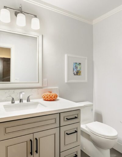 Modern bathroom with a white vanity, mirror, and three geometric vases. Features a toilet, wall art, and overhead lighting in a neutral-toned space.