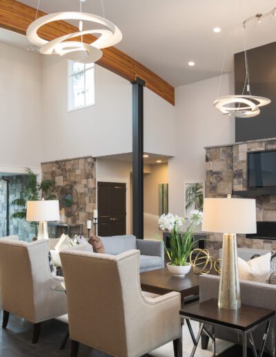 Modern living room with high ceiling, stone accent wall, large windows, two beige armchairs, a gray sofa, coffee table with lamps, and a wall-mounted TV above a fireplace.