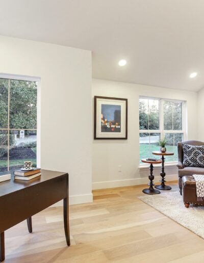 A modern study with a desk and lamp on the left, a cozy sitting area with two chairs, a small table, and a bookshelf on the right. Large windows provide natural light and a view of the garden.