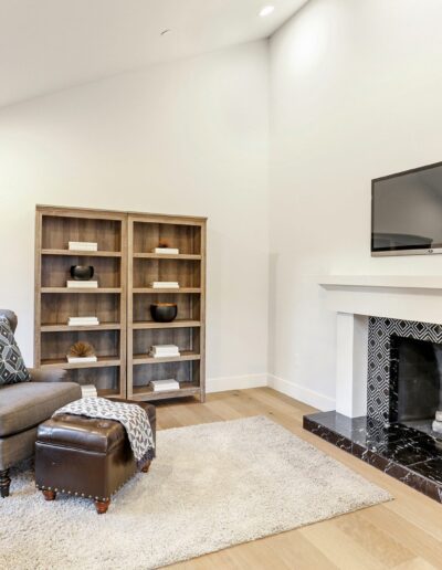 A cozy living room with an armchair, ottoman, small side tables, a bookshelf, and a TV above a decorative fireplace. A potted plant and a window are on the left.