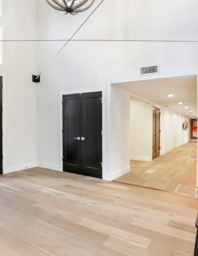 Spacious, bright hallway with light wood flooring, a black door, double black doors, and a bench with patterned cushions. A hallway extends to the right.