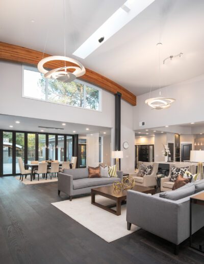 Modern open-concept living and dining area with large windows, gray sofas, overhead lights, and a view of the kitchen.