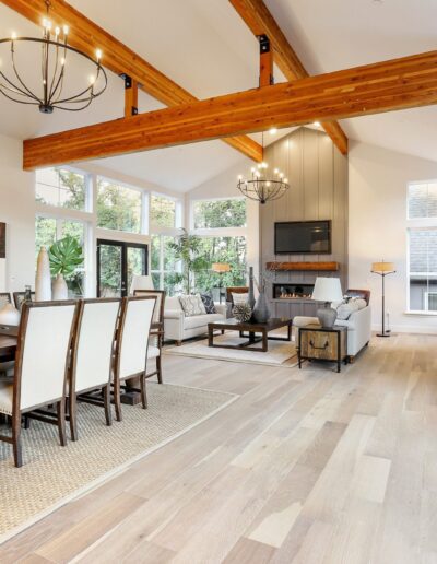 Open-concept living room with wooden beams, modern chandeliers, dining area, and a seating area with a fireplace and TV. Large windows provide natural light.