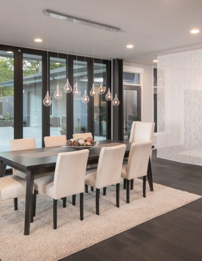 Modern dining room with a large table, eight white chairs, and pendant lighting. Glass doors open to a patio. Dark wood flooring and neutral decor create a minimalist aesthetic.