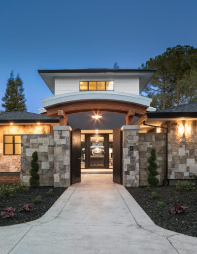 Modern house exterior with stone and wood accents, large windows, and a central entrance. Surrounded by landscaped garden and trees, under a twilight sky.