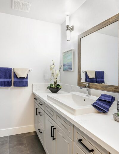 Bathroom with a white vanity, large mirror, dual sinks, and blue towels. Modern decor features a metal tree sculpture and framed art on white walls.