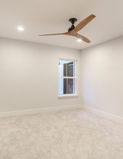 Empty room with light gray carpet, white walls, a window, black sliding door, and a modern wooden ceiling fan.