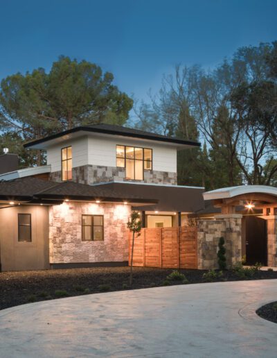 Modern house with stone and stucco exterior, surrounded by trees, featuring warm lighting and a curved driveway.