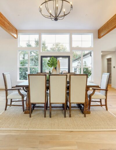 A dining area with six chairs around a wooden table, a chandelier above, and large windows. To the right is a modern kitchen, and to the left is a living area with a couch.