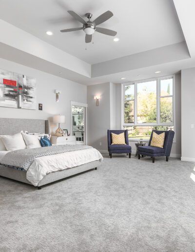 Spacious modern bedroom with a large bed, gray carpet, two chairs, ceiling fan, and large windows letting in natural light.