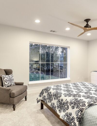 Cozy bedroom with a gray armchair, bed with patterned bedding, large window, ceiling fan, and wall art.