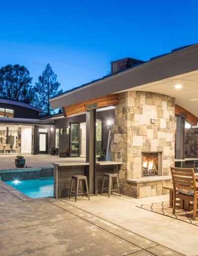 Modern outdoor patio with a stone fireplace, dining table, stools, and pool, set against an illuminated contemporary house in the evening.