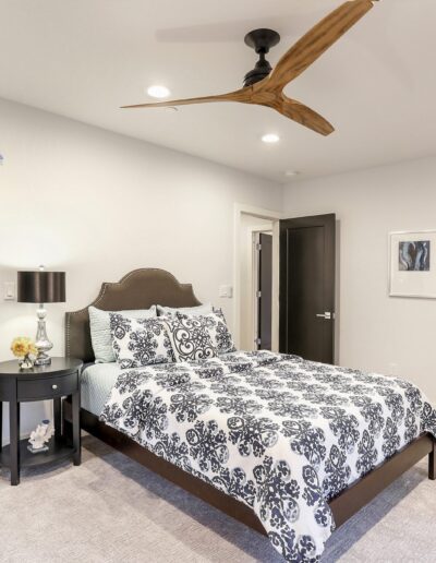 A modern bedroom with a patterned bedspread, two wall art pieces, an armchair, a ceiling fan, and an open walk-in closet. The room has neutral colors and simple decor.