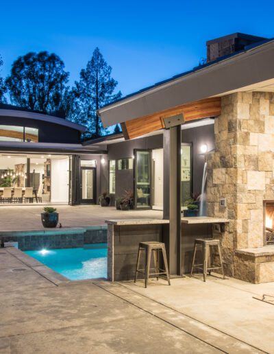 A modern outdoor patio with a stone fireplace, seating area, and a small pool, lit by exterior lights at dusk.