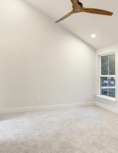 Empty room with light gray carpet, white walls, large window, and wooden ceiling fan.