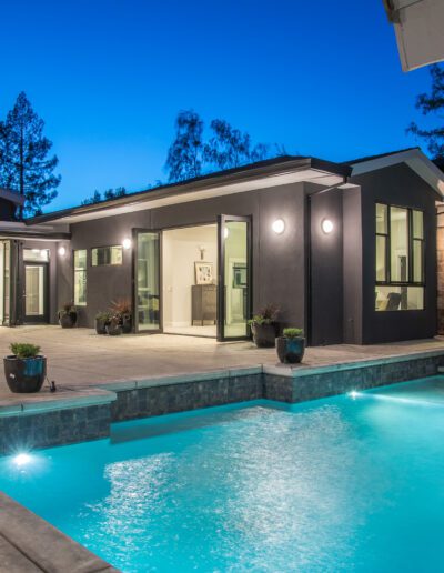 Modern house with large windows, an open dining area, and a lit swimming pool featuring a waterfall at dusk.