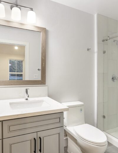 Modern bathroom with a large mirror, single sink vanity, toilet, and a glass-enclosed shower. The walls are white, and the room is illuminated by three overhead lights.